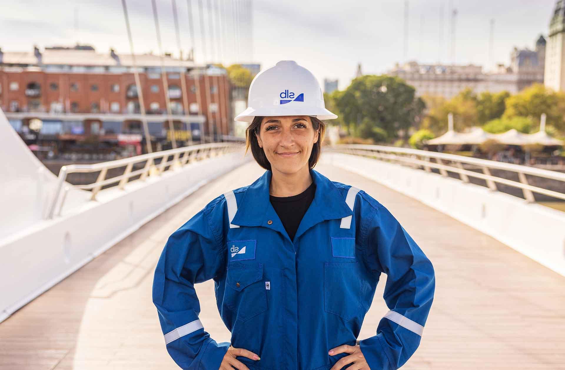 Female 全国快3信誉最好的老平台 employee in blue coveralls standing on a bridge with a city in the background